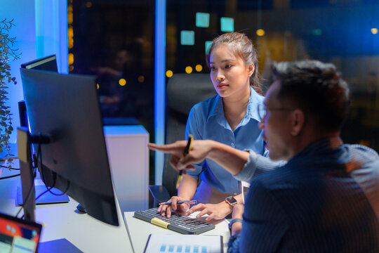 Asian woman project manager and developer pointing at computer screen explaining about code algorithm and discussing to a new male software developer at desk in office. Developer team collaborating.