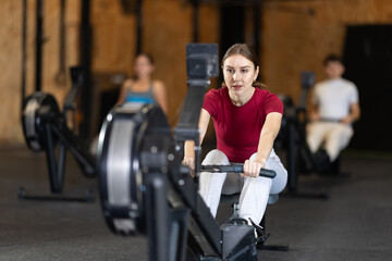Girl hardly performs exercises on rowing machine during fitness class. Gym and fitness equipment,...