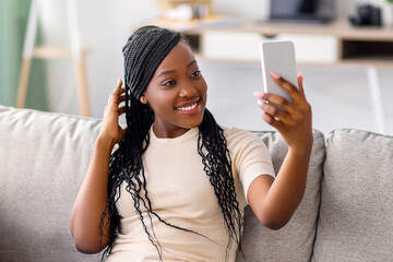 Cute young black lady with stylish dreadlocks taking selfie on smartphone for social media or friends, positive african american woman having fun at home, using mobile phone, copy space
