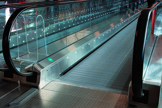 Almost empty moving walkway in an international airport corridor. Reflection of few people dragging suitcases. Futuristic design of glass railings.