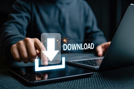 Person s hand interacting with a tablet displaying a glowing download icon and the word download next to a laptop