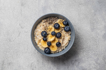 Oatmeal porridge in bowl with milk, blueberries, banana, chia seeds and honey on a grey background. Healthy breakfast homemade food. Home cooking. Top view, selective focus.