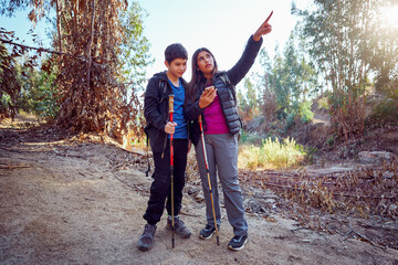 Two young latin Americans hikers using trekking poles and consulting navigation app on smartphone...