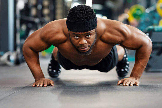 Push-ups, press-ups workout. Muscular african american shirtless guy athlete building up muscles, black man training on floor at modern gym, doing push-ups and looking at camera