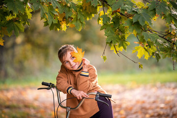 A young girl on a bicycle covering her eye with a vibrant orange autumn leaf