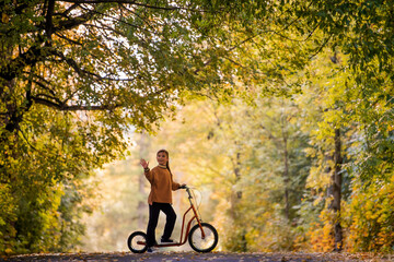 A girl on a scooter waves her hand on an autumn road, outdoor activity.