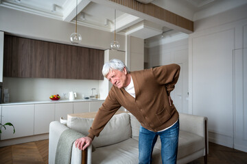 Lonely retiree with backache leans on sofa for support due to severe lumbar pain. Old male suffer from osteoporosis, pinched nerves, muscle atrophy, abandoned elderly, self care difficulties, hernia