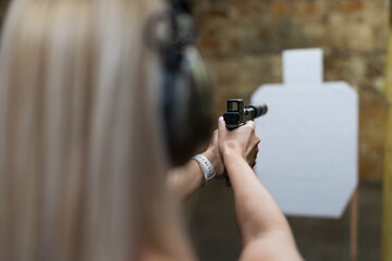 A woman in protective headphones focusedly aims a pistol with a collimator sight and a suppressor...
