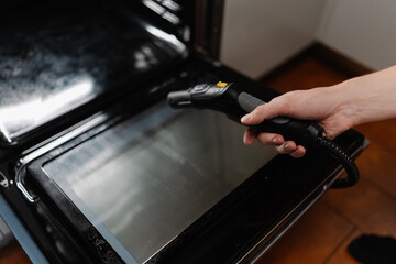 Close-up cropped shot of unrecognizable man steam cleaning kitchen oven surface, targeting stubborn...