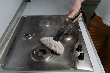 Top view of housewife cleaning dirty gas stove using steam cleaner and cloth, removing grease and...