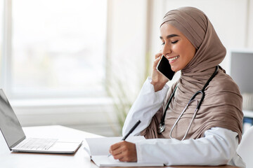 Smiling young lady in hijab doctor talking on mobile phone and taking notes, sitting at workdesk in...