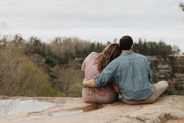 Fototapeta premium Man and woman sitting side by side on a rocky cliff edge overlooking a forest valley with arm around in a serene romantic outdoor moment
