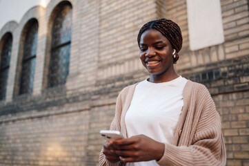 Smiling young woman using smartphone and wearing wireless earbuds outdoors