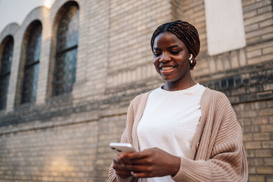 Smiling young woman using smartphone and wearing wireless earbuds outdoors