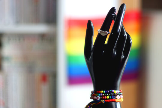 Hand figurine with colorful rainbow bracelets and heart shaped ring. Pride flag in the background. Pride month concept, selective focus.