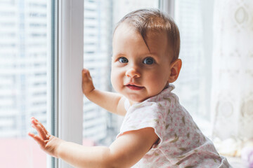 Cute baby girl infant looking out the window with curious expression, dressed in light clothing in a bright room, concept of childhood and innocence