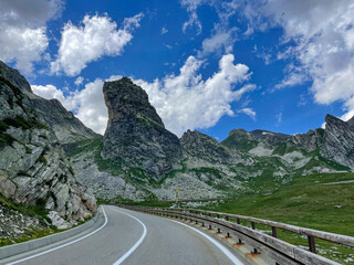 Smooth winding asphalt road cuts through dramatic alpine landscape under cloudy blue sky. Towering...