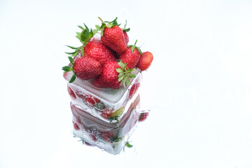 Bright red strawberries stacked on a transparent ice cube, reflected on a glossy white surface....
