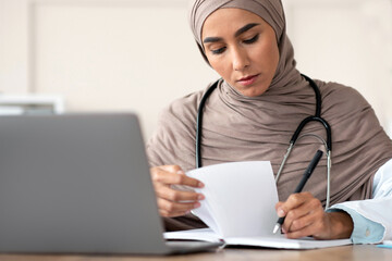 Concentrated young woman doctor in hijab taking notes in hospital, sitting in front of laptop, using notebook and pen, copy space. Female therapist in head scarf planning her working day