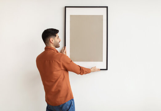 Modern Home Interior And Domestic Decor. Smiling young man hanging painting, putting photo picture frame on the wall. Casual guy holding showing empty poster, design mockup, blank free copy space