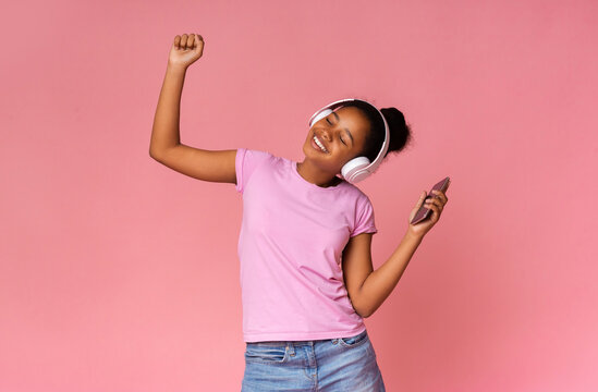 Energetic african teen dancing, listening to favorite music with wireless headphones, pink studio background