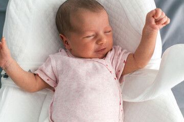 Newborn baby in pink onesie with red fruit print lying in a white baby seat with eyes closed and arms raised, concept of childhood and care 