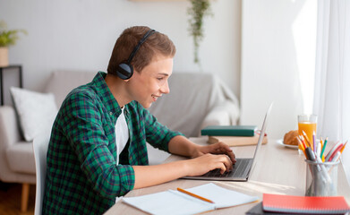 Cheerful schooler with headset typing on laptop at home, having online test, empty space