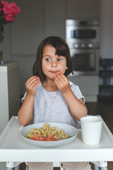 Little cute girl toddler chewing food with puffed cheeks and playful expression, sitting at the table with pasta and salmon in cozy kitchen, concept of childhood, vertical portrait