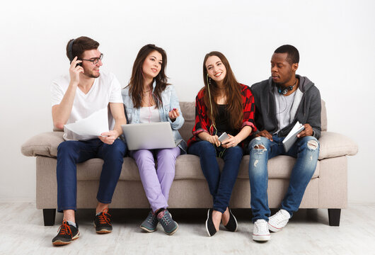 Young diverse students, people listen to music with gadgets, sitting together on couch indoors. Modern education.