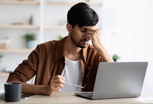 Tired indian man sitting in front of laptop at office, touching his head, suffering from burnout or financial crisis, young guy freelancer having difficulties with job, lack of clients, copy space