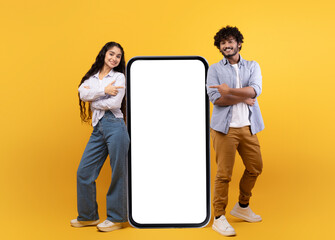 Young indian spouses posing near huge smartphone with mockup, promoting mobile app or website,...