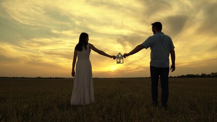 Family hands are holding paper house at sunset, sun is shining through window. Symbol of house, happiness. Concept of building house for family. Dream to buy house. Home for children and parents