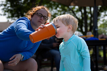 Young child getting a drink from a large tumbler with help from his mother.