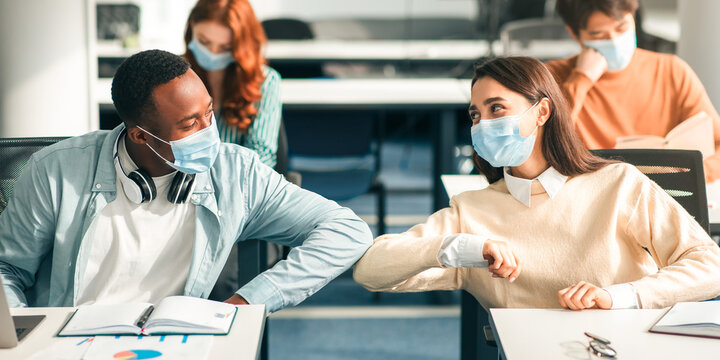 Young multiethnic classmates wearing protective disposable surgical masks greeting each other and bumping elbow at classroom. Smiling woman and black guy avoid touching, studying, sitting at desk