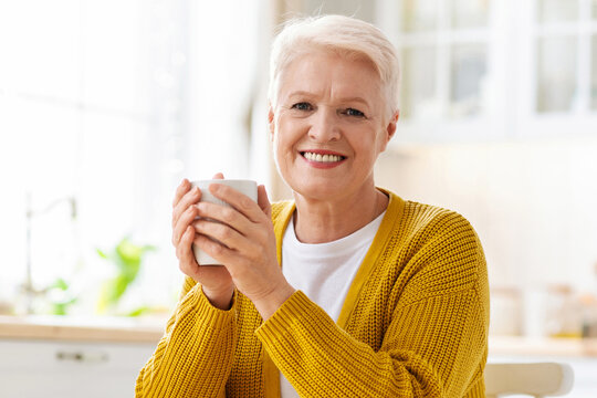 Cheerful elderly lady drinking coffee with croissant, sitting in kitchen at home, having snack, copy space. Attractive senior woman enjoying cup of herbal tea with home baked pastry - Powered by Adobe