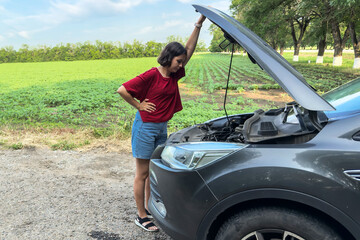 Young woman in red shirt and denim shorts standing on roadside, examining car engine with hood open, looking concerned during road trip in rural area, transportation concept