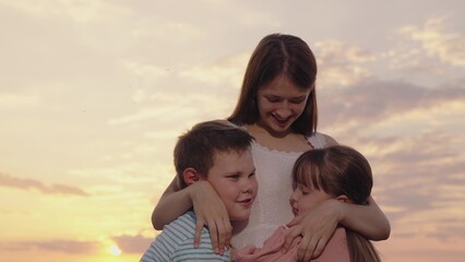 Family, young mother hugging children, boy girl in park against sky. Happy children daughter son hugging mommy in summer park. Carefree childhood, joyful hugs of mother child. Happy family in nature