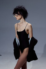 Slim woman wearing black bodysuit and sunglasses poses confidently with jacket on arms against...