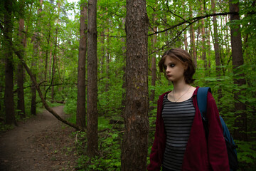 Naklejka premium Beauty face young woman with serious thinking look hiking among green pine trees on the path in the forest with blue backpack on shoulder. Girl walking in woods, in fashion clothes Emotion portrait