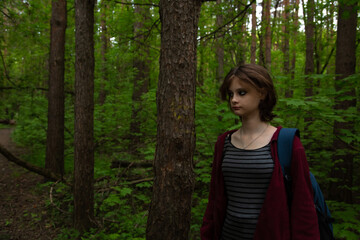 Naklejka premium Beauty face young woman with serious thinking look hiking among green pine trees on the path in the forest with blue backpack on shoulder. Girl walking in woods, in fashion clothes Emotion portrait