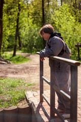 A person with short hair leans against a railing, gazing into the distance. They're wearing a gray jacket and pants. The background features a sunlit forest with a dirt path