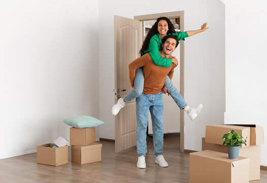 Celebrating Moving Day. Happy couple celebrating relocation to their own home, cheerful guy giving lady piggyback ride, excited young millennial family of two having fun standing in new flat - Powered by Adobe