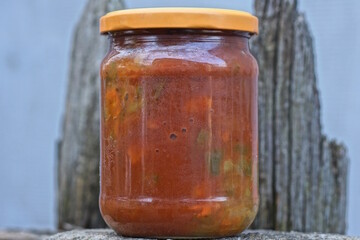 one closed glass jar with red lecho from canned pepper stands on a gray table on an autumn street