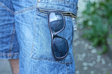 one black plastic sunglasses hanging in pocket blue cotton jeans on leg on summer street