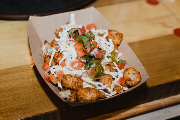 Loaded tater tots topped with shredded cheese, pico de gallo and jalapeños in a paper tray on...