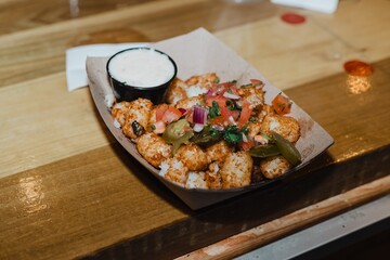 Paper tray of loaded tater tots topped with pico de gallo and pickled jalapeños beside a cup of...