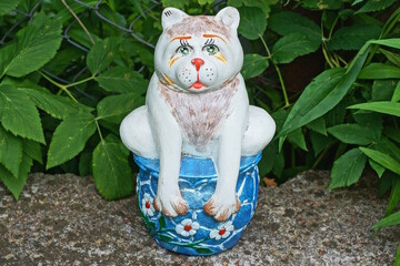 one large ceramic colored bank piggy bank with a figure of a cat stands on a gray table against the backdrop of green vegetation on a summer street