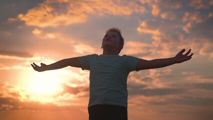 Child plays in park against sky. Little boy prays against background of sky and sun. Child raised his hands to sky in park at sunset, true faith. Religion and god, childhood dreams. Happy family