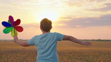 Family vacation in nature, sky, Happy kid plays with toy pinwheel outdoors in spring park in sunshine. Childhood, children. Little boy runs with toy wind turbine in his hand on summer field at sunset.