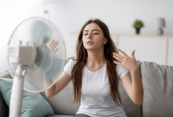 Sad young european woman suffers from unbearably too hot weather, catches cold air from fan in...